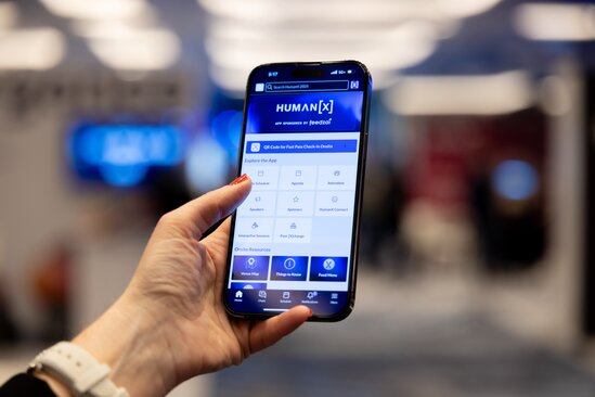 a hand holds a phone which is open on the humanx event app. the showfloor is blurred in the background